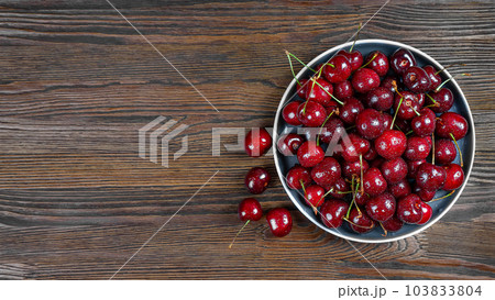 Fresh red ripe sweet cherry with water drops on plate on brown wooden background. Fresh red ripe sweet cherry with water drops on plate on brown wooden background. 103833804
