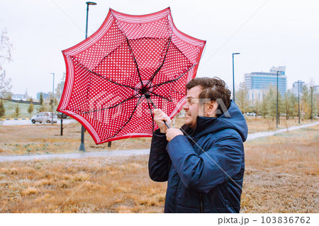 Shocked European white young male man fool around with umbrella in city park, strong storm wind, rain and bad weather. Wind broke umbrella. Autumn waist up lifestyle portrait. 103836762