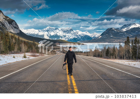 Young asian photographer man walking on the highway with rocky mountains and frozen lake in winter Young asian photographer man walking on the highway with rocky mountains and frozen lake in winter 103837413
