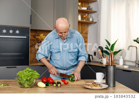 Retired senior man cooking from green salat, pepper and tomatoes. Man standing in kitchen interior. Man wearing jeans and shirt. Retired senior man cooking from green salat, pepper and tomatoes. Man standing in kitchen interior. Man wearing jeans and shirt. 103843532