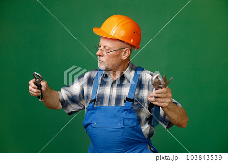 Studio portrait of a man isolated on green background. Seniour man worker in a blue plaid shirt and blue overall holds a straight pipe wrench or spanner. Man wearing eyeglasses and orange helmet. 103843539