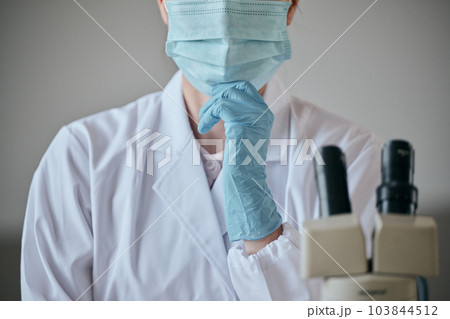 デスクで顎に手を当てて考え事をする白衣の若い女性 103844512