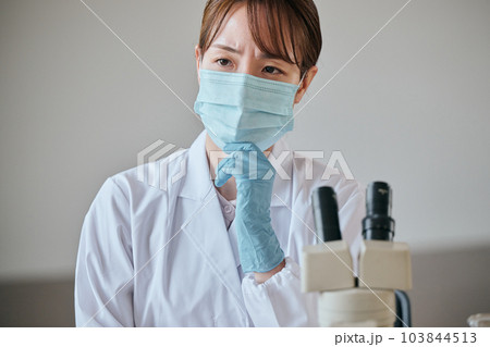 デスクで顎に手を当てて考え事をする白衣の若い女性 103844513