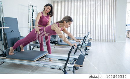 Asian woman doing plank on a reformer under the control of a trainer. Asian woman doing plank on a reformer under the control of a trainer. 103846552