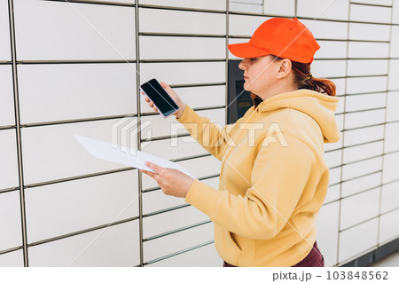 Young woman in red hat picks up parcel from automatic post office machine, Courier standing with phone and small box. Concept of fast delivery to automatic self lockers Young woman in red hat picks up parcel from automatic post office machine, Courier standing with phone and small box. Concept of fast delivery to automatic self lockers 103848562