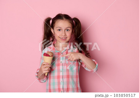 Adorable happy little child girl with two ponytails, wearing checkered shirt, smiling looking at camera, showing a waffle cone with delicious ice cream over isolated pink background. Food. Summer Adorable happy little child girl with two ponytails, wearing checkered shirt, smiling looking at camera, showing a waffle cone with delicious ice cream over isolated pink background. Food. Summer 103850517