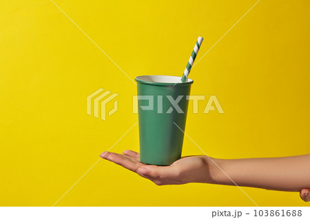 A green disposable paper cup with a paper straw placed in a woman's hand on a yellow background. Minimal scene for advertising and branding drinking product. Copy space A green disposable paper cup with a paper straw placed in a woman's hand on a yellow background. Minimal scene for advertising and branding drinking product. Copy space 103861688