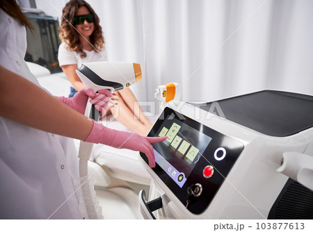 Close-up view hands of beautician in pink gloves, who holding laser sensor with one hand and adjusting laser device on scoreboard with the other. Satisfied client in the blurred background. 103877613