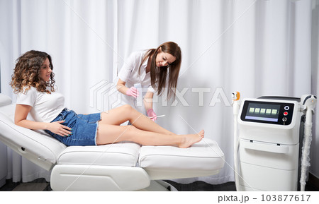 Smiling cosmetologist in sterile gloves applying gel on female leg before epilation. Young woman lying on daybed near laser diode epilation machine while having hair removal procedure in clinic. 103877617