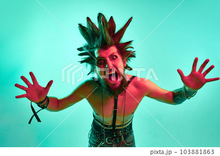 Portrait of young crazy looking man, punk with expressive hairstyle posing shirtless against blue studio background in neon light Portrait of young crazy looking man, punk with expressive hairstyle posing shirtless against blue studio background in neon light 103881863