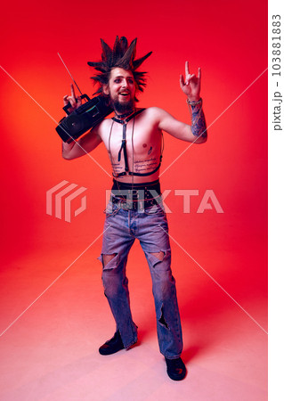 Young man, punk in extraordinary clothes and hair listening to music with retro player against red studio background in neon light 103881883