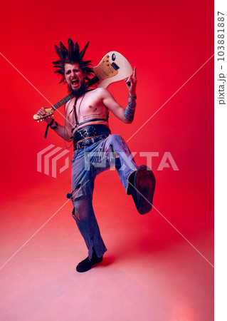 Portrait of creative young man, punk, musician posing with electric guitar against red studio background in neon light Portrait of creative young man, punk, musician posing with electric guitar against red studio background in neon light 103881887