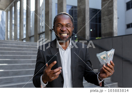 Mature African American businessman is happy about the successful results of financial activity, man is holding in his hands a smartphone and cash American money dollars, outside the office building. 103883814