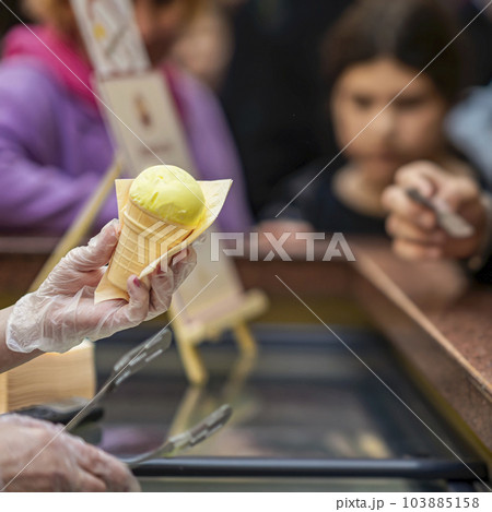 Hand Of Vendor Holding fruit Ice Cream Cone, background of counter with sweet dessert. Real scene in store Hand Of Vendor Holding fruit Ice Cream Cone, background of counter with sweet dessert. Real scene in store 103885158