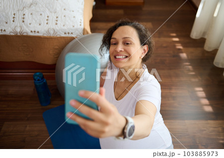 Beautiful pregnant woman sitting on blue exercise mat at home, smiling broadly taking selfie on smart mobile phone, sharing her happy moments of her pregnancy and maternity lifestyle in social media Beautiful pregnant woman sitting on blue exercise mat at home, smiling broadly taking selfie on smart mobile phone, sharing her happy moments of her pregnancy and maternity lifestyle in social media 103885973