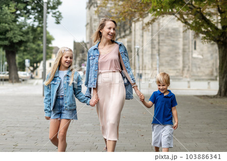 Young adult beautiful mom enjoy having fun walking with two kids in european city street at summer day. Happy parenthood family concept. Mother care resonsibility. People go outside in Germany 103886341