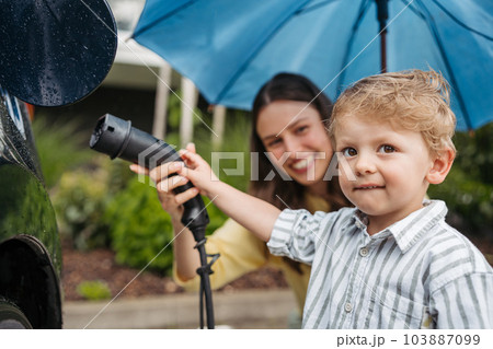 Close up of smiling mother and her little son charging their electric car on a rainy day, standing under umbrella. Close up of smiling mother and her little son charging their electric car on a rainy day, standing under umbrella. 103887099