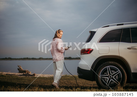 Young woman holding power supply cable from her electric car, prepared for charging it sustainable and economic transportation concept. Young woman holding power supply cable from her electric car, prepared for charging it sustainable and economic transportation concept. 103887145