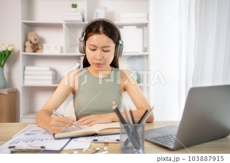 Beautiful young asian woman watching live video or video call of teacher teaching on laptop in her home, Take notes of important conversations and messages during the teacher's teaching. 103887915