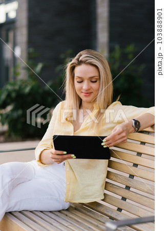Beautiful woman sits on a bench in the summer in the city and makes online purchases using a smartphone or communicates via chat Beautiful woman sits on a bench in the summer in the city and makes online purchases using a smartphone or communicates via chat 103888901