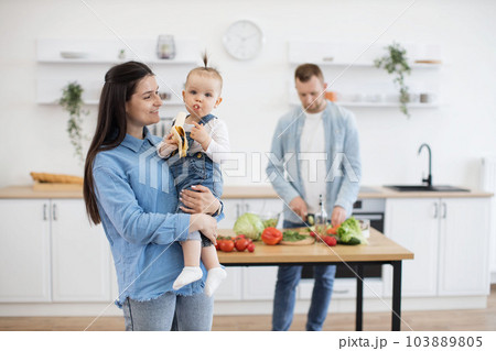 Baby eating banana on mommy's hands while dad making dinner 103889805