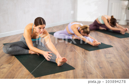 Yoga class of relaxed sports women performing Head to Knee Forward Bend Pose, or Janu Sirsasana after practicing together in gym Yoga class of relaxed sports women performing Head to Knee Forward Bend Pose, or Janu Sirsasana after practicing together in gym 103894278
