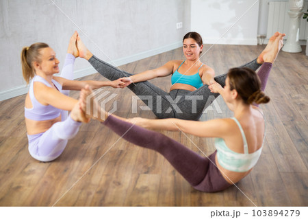 Smiling young women sitting on flour, holding hands and touching each others feet after training together in fitness center Smiling young women sitting on flour, holding hands and touching each others feet after training together in fitness center 103894279