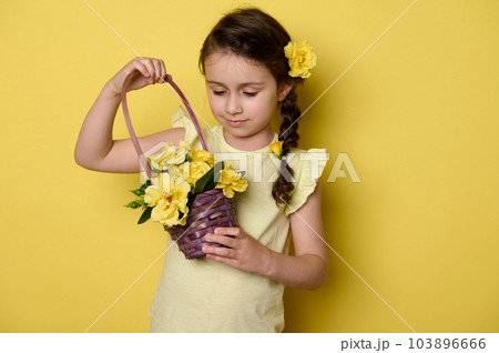 Adorable little child girl with rose flowers in hairstyle, wearing a yellow summer dress, holding a purple wicker basket full of fresh yellow roses, isolated yellow background. Happy Children's Day Adorable little child girl with rose flowers in hairstyle, wearing a yellow summer dress, holding a purple wicker basket full of fresh yellow roses, isolated yellow background. Happy Children's Day 103896666