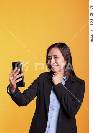 Cheerful asian woman talking with remote friend during videocall meeting conference, having remote discussion during shoot break in studio. Adult in formal suit standing over yellow background Cheerful asian woman talking with remote friend during videocall meeting conference, having remote discussion during shoot break in studio. Adult in formal suit standing over yellow background 103900570