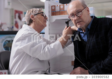 Old man getting ear examination procedure in pharmacy store, checking for elderly hearing loss problem. Woman pharmacist examining senior customer with otoscope medical tool in apothecary 103900582