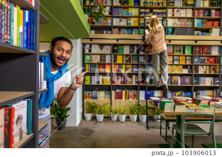 Young dark-skinned man hiding behind the bookshelves Young dark-skinned man hiding behind the bookshelves 103906013