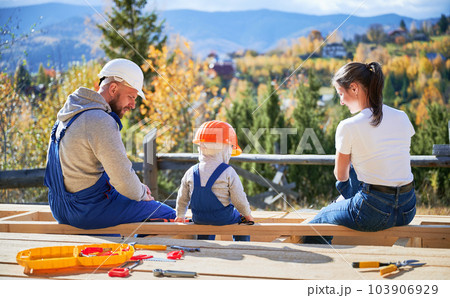 Back view of father, mother and son building wooden frame house. Toddler boy, daddy and woman having a rest on construction site. Guys wearing helmet and blue overalls. Carpentry, family concept. Back view of father, mother and son building wooden frame house. Toddler boy, daddy and woman having a rest on construction site. Guys wearing helmet and blue overalls. Carpentry, family concept. 103906929