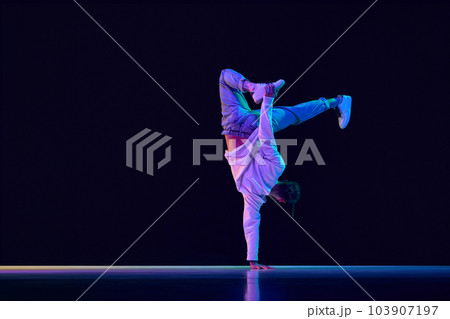 Youth culture. Young man in casual clothes making performance, dancing breakdance against black studio background in neon light Youth culture. Young man in casual clothes making performance, dancing breakdance against black studio background in neon light 103907197