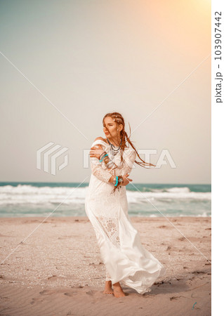 woman sea white dress. Model in boho style in a white long dress and silver jewelry on the beach. Her hair is braided, and there are many bracelets on her arms. 103907442