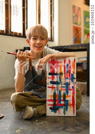 Full length view of young gay male artist sitting on the floor in art studio lit by sunlight Full length view of young gay male artist sitting on the floor in art studio lit by sunlight 103908152