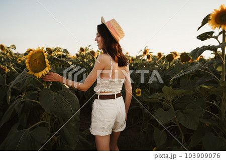 Smiling woman wearing a hat standing in a field of sunflowers in sunset Smiling woman wearing a hat standing in a field of sunflowers in sunset 103909076