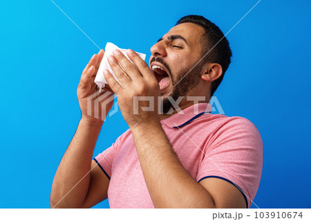 Young man in pink shirt with allergy or cold, blowing his nose in a tissue against blue background Young man in pink shirt with allergy or cold, blowing his nose in a tissue against blue background 103910674
