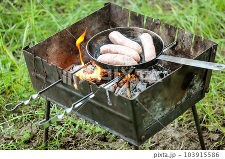 Grilling row sausages. Appetizing sausage, wieners, meats fried on black griddle on picnic fire view from above. Cooking outdoors in the park woods. Barbecue on coals, street grill on open fire flatly 103915586