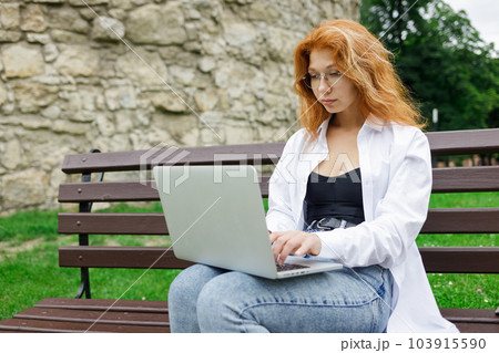 Young female student using her laptop sitting on the bench in park 103915590