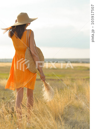 Beauty romantic girl outdoors. Rear view of a beautiful girl dressed in a casual orange dress with a straw hat and a straw bag in her hands in a field in the sunlight. Blows long hair. Autumn. Shine 103917507