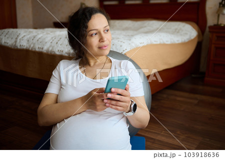 Multi-ethnic thoughtful pretty gravid female, pregnant woman, expectant mother holding mobile phone, checking new application, shopping online, dreamily looking aside, sitting on yoga mat at home 103918636