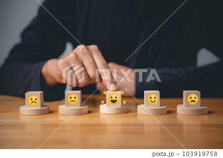 Man's hand chooses an emotion smiley face on a wooden cube to rate excellent business services. Satisfaction survey and feedback concept. 5-star satisfaction is guaranteed. 103919758