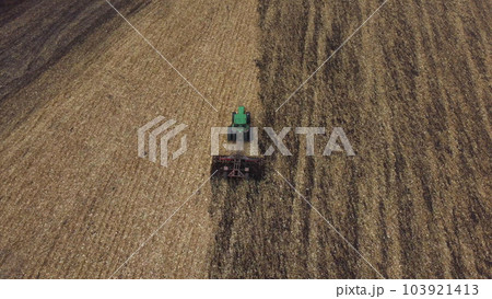 Tractor plowing the ground. Flying over green tractor that plows up ground in yellow field after harvesting wheat on autumn day. Tractor digging land. Agricultural work on field. Aerial drone view 103921413