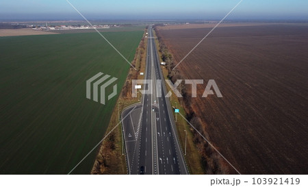 Beautiful panoramic landscape. Black paved highway with white markings,many driving passing cars, large agricultural fields with green agricultural crops and plowed black soil on a sunny autumn day. Beautiful panoramic landscape. Black paved highway with white markings,many driving passing cars, large agricultural fields with green agricultural crops and plowed black soil on a sunny autumn day. 103921419