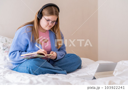 Attentive teen girl listening to podcasts in headphones making notes in diary teenager sitting on bed near modern tablet learning new information at home Attentive teen girl listening to podcasts in headphones making notes in diary teenager sitting on bed near modern tablet learning new information at home 103922241