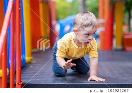 Curious preschooler with Down syndrome sitting on colorful climber at modern playground in residential complex blond kid enjoying outdoor activities in city 103922267