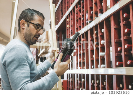 A white Caucasian man holding a red wine bottle, reading the label, in a specialist wine shop A white Caucasian man holding a red wine bottle, reading the label, in a specialist wine shop 103922616