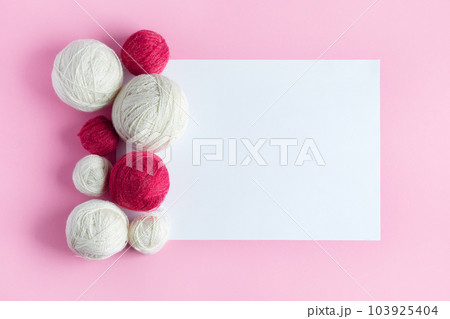 Magenta and white balls of knitting thread with a white sheet of paper for text. Top view, flat lay, work space on pink background. Magenta and white balls of knitting thread with a white sheet of paper for text. Top view, flat lay, work space on pink background. 103925404