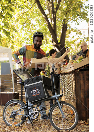 Organic locally grown fruits and vegetables delivery at farmers market, courier putting bio organic produce in thermal backpack and picking up order. Delivering food order fron local natural stand. Organic locally grown fruits and vegetables delivery at farmers market, courier putting bio organic produce in thermal backpack and picking up order. Delivering food order fron local natural stand. 103926898