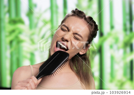 Attractive woman doing hairstyle and using hairbrush as microphone. Young beautiful caucasian lady listening to music, pretending to sing in comb and brushing blonde hair Attractive woman doing hairstyle and using hairbrush as microphone. Young beautiful caucasian lady listening to music, pretending to sing in comb and brushing blonde hair 103926914